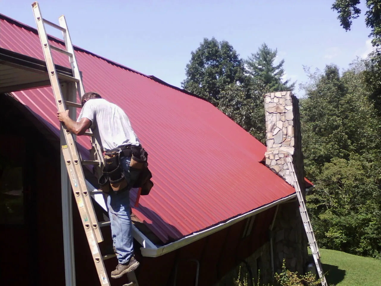 Roofer on a ladder beside a red metal roof and stone chimney — Soffit & Fascia Repair by Roofing Brothers in Saint James City